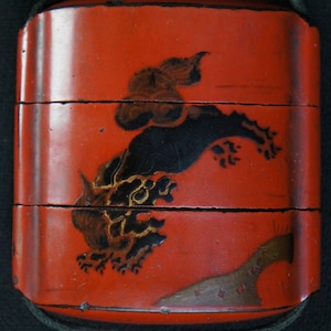 May include: A three-tiered red lacquer box with a black and gold dragon design. The box has a black cord and is likely used for carrying food or small items. The dragon is painted on the front.