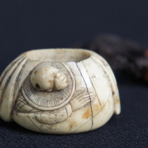 Könnte beinhalten: Ein cremefarbener, kuppelförmiger Ring mit einem komplizierten geschnitzten Relief eines Schädels, der auf einer kreisförmigen Basis mit strahlenden Linien ruht. Die Oberfläche weist eine natürliche Alterung mit feinen Rissen und dunklen Details auf.