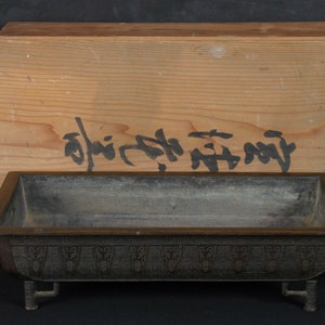 May include: A rectangular, dark grey, metal planter with a decorative border and four short legs. The planter is sitting on top of a wooden box with Japanese writing on the lid.