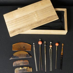 May include: A collection of antique hair accessories, including combs and hairpins, displayed on a black surface. The set includes various sizes and styles, with decorative elements and a wooden box.