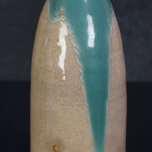 May include: A ceramic bottle with a beige body and a teal glaze that drips down the front. The rim is a darker brown. The bottle has a textured, crackled surface.