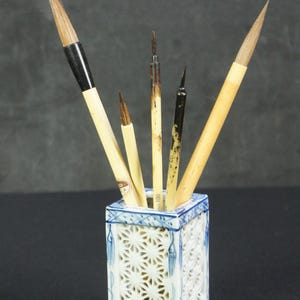 May include: A square ceramic brush holder with blue and white lattice patterns containing four calligraphy brushes of varying sizes. The brushes feature light-colored wooden handles and dark bristles, with one brush handle clearly marked with the text No 100.