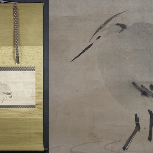 May include: A traditional Japanese hanging scroll featuring a black ink painting of a bird on a light brown background. The scroll is mounted on a gold fabric with a repeating pattern of small circles.