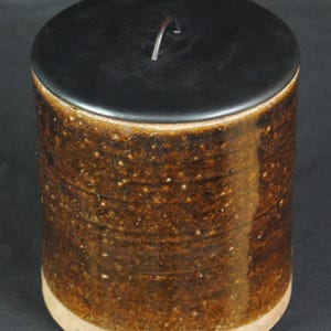 May include: A cylindrical ceramic canister with a dark brown, speckled, glazed finish and a smooth, unglazed light tan base. The container features a flat, circular black lid with a small, thin, dark metal wire loop handle centered on top.