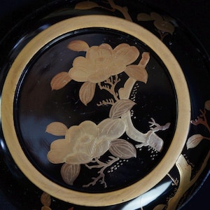 Puede incluir: Un plato decorativo negro con borde dorado y diseño floral. El plato presenta flores y hojas doradas, creando un contraste con el fondo oscuro. El diseño es intrincado y detallado.