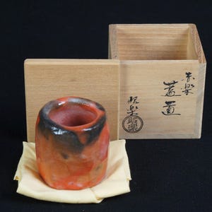May include: A handmade, cylindrical ceramic cup with a reddish-orange glaze and black accents, resting on a beige cloth. The cup is next to a wooden box with Japanese characters and a stamp.