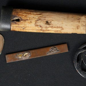 May include: A selection of antique Japanese sword fittings, featuring a wooden scabbard, two oval tsuba, a rectangular piece with a landscape design, and a decorative guard. The components are in shades of brown, silver, and gold.