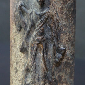 May include: A detailed carving of a figure in a flowing robe, with a crown and a hand holding a fan. The carving is on a weathered, grey surface.