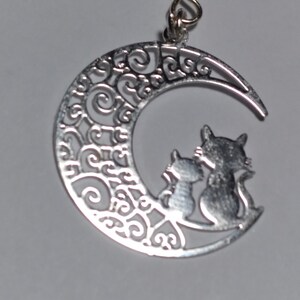 May include: Silver-toned cat-themed earring. The earring features a crescent moon design with intricate scrollwork and two cat silhouettes. The earring has a hook closure.