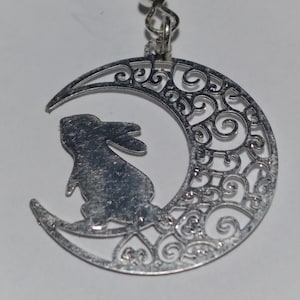 May include: Silver tone earrings featuring a crescent moon design with a detailed filigree pattern and a rabbit silhouette inside the moon.