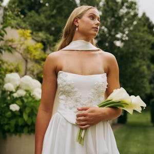 May include: A white strapless wedding dress with a fitted bodice and a flowing skirt. The bodice is adorned with delicate beading and lace. The bride is holding a bouquet of white calla lilies.