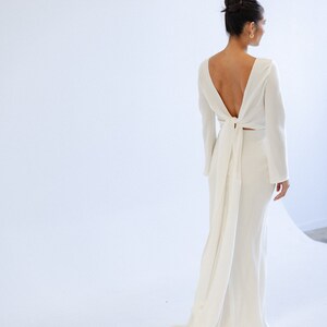 May include: A floor-length ivory crepe wedding gown with long sleeves and an open back. The bodice features a unique tie detail at the back, creating a flowing train. The skirt is form-fitting, and the overall design is minimalist and elegant.