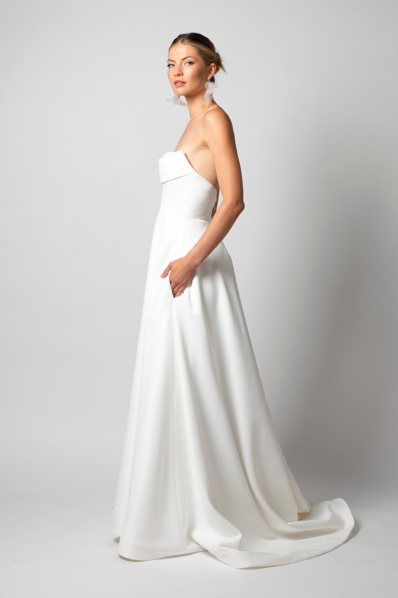 May include: A white strapless wedding dress with a simple, flowing skirt and a pocket on the left side.