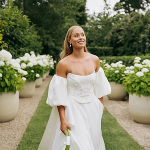 May include: A bride wearing a white off-the-shoulder wedding dress with a fitted bodice and a flowing skirt. The dress has puff sleeves and is decorated with delicate floral embroidery. The bride is holding a bouquet of white calla lilies and is walking down a path lined with white flowers and potted plants.