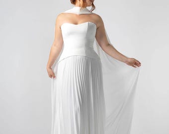 Puede incluir: Un vestido de novia blanco sin tirantes con falda plisada y chal a juego. El vestido presenta un corpiño ajustado y una falda fluida. La modelo lleva el vestido en un estudio con fondo blanco.