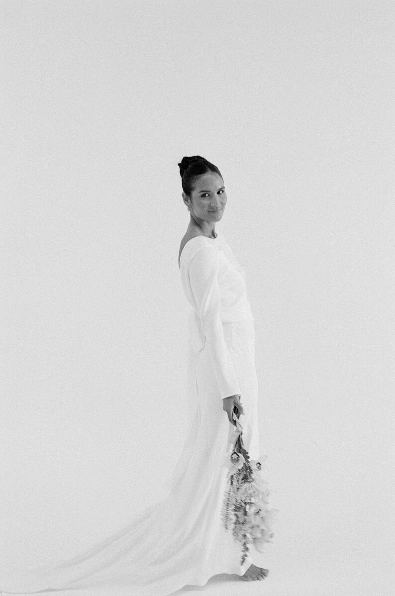 May include: A long-sleeved, form-fitting, white bridal gown with a low back and a flowing train. The gown has a minimalist design and is shown on a model holding a bouquet of light-colored flowers.