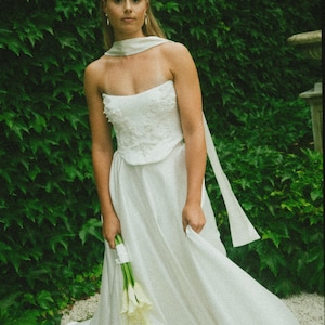 May include: A white strapless wedding dress with a fitted bodice and a flowing skirt. The dress has a white sash tied around the neck. The bride is holding a bouquet of white calla lilies.