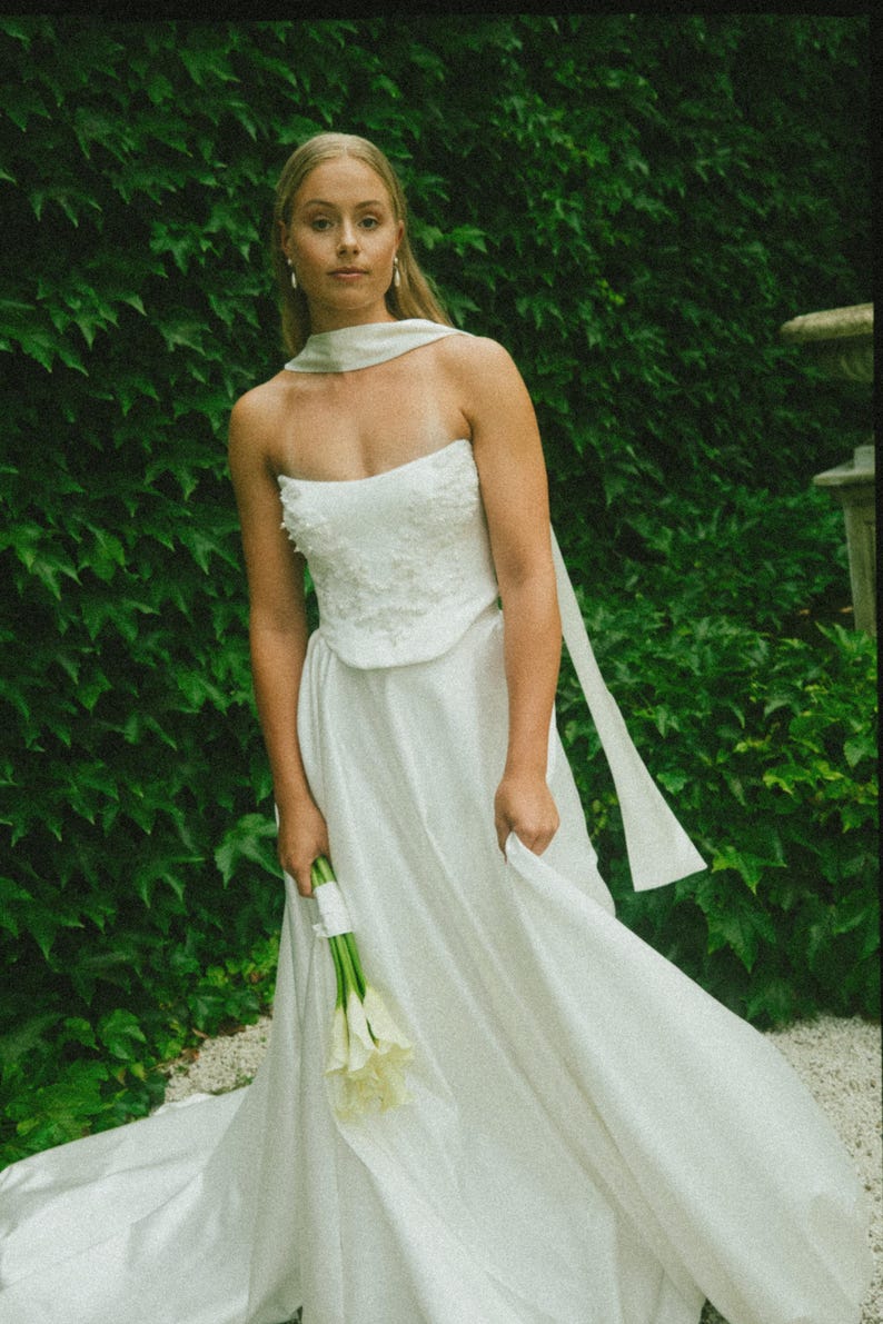 May include: A white strapless wedding dress with a fitted bodice and a flowing skirt. The dress has a white sash tied around the neck. The bride is holding a bouquet of white calla lilies.