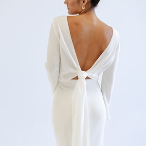 May include: A white silk maxi dress with long sleeves and a unique open back detail. The back features a self-tie closure that creates a bow. The dress has a form-fitting silhouette and a floor-length skirt. The fabric appears smooth and luxurious.