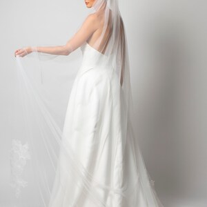 May include: A white bridal veil with delicate floral lace detailing. The veil is made of soft, sheer tulle and has a long, flowing design.