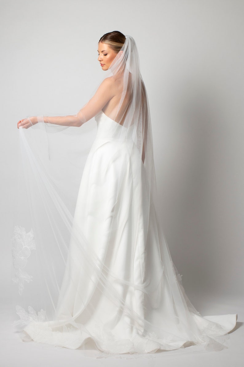 May include: A white bridal veil with delicate floral lace detailing. The veil is made of soft, sheer tulle and has a long, flowing design.
