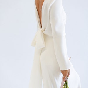 May include: A white long-sleeve wedding gown with a low back and a bow detail. The dress is made of a sleek, flowing fabric and has a form-fitting silhouette.  The long sleeves add elegance, and the back features a unique, stylish tie. The gown is paired with a bouquet of orchids and dahlias.