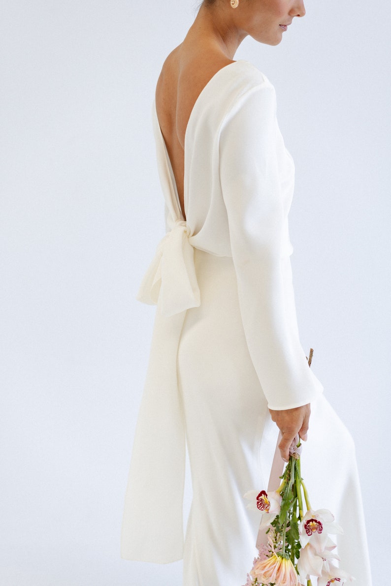 May include: A white long-sleeve wedding gown with a low back and a bow detail. The dress is made of a sleek, flowing fabric and has a form-fitting silhouette.  The long sleeves add elegance, and the back features a unique, stylish tie. The gown is paired with a bouquet of orchids and dahlias.
