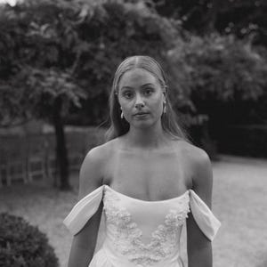 May include: A woman wearing a white off-the-shoulder gown with a beaded bodice. The gown has a simple, elegant design and is made of a soft, flowing fabric.