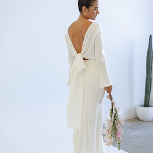May include: A white long-sleeved maxi dress with a low back and a bow tie detail. The dress is made of a smooth, flowing fabric and has a fitted silhouette. The dress is shown on a mannequin against a white background. The dress has a train.