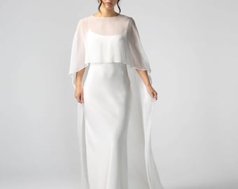 Puede incluir: Un vestido de novia blanco con una capa transparente y fluida. El vestido tiene un diseño simple y elegante con un corpiño ajustado y una falda larga y fluida. La capa añade un toque de belleza etérea al vestido de novia.