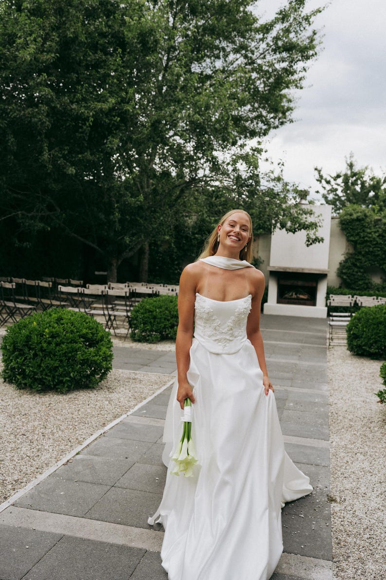 May include: A bride in a white strapless wedding dress with a white sash and floral embellishments. She is holding a bouquet of white calla lilies and smiling.