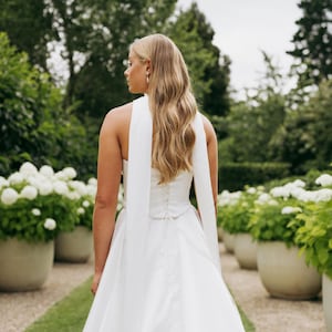 May include: A white halter neck wedding dress with a long train. The dress has a fitted bodice and a flowing skirt. The dress is accessorized with a long white sash that ties at the back.