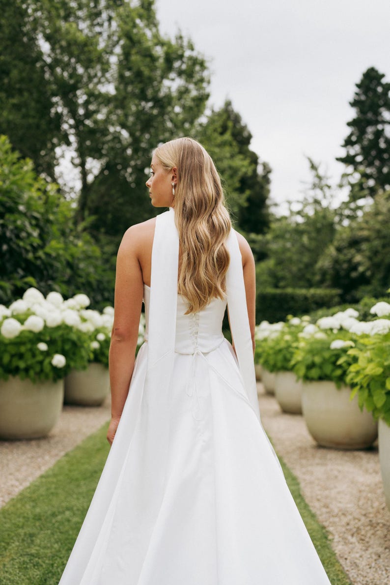 May include: A white halter neck wedding dress with a long train. The dress has a fitted bodice and a flowing skirt. The dress is accessorized with a long white sash that ties at the back.