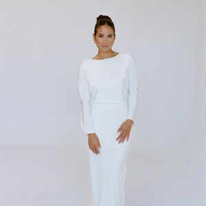 May include: A floor-length, ivory satin wedding dress with long sleeves and a fitted silhouette. The bodice is a cropped style, creating a defined waistline. The skirt flows smoothly to the floor, with a subtle train. The dress is simple and elegant, suitable for a modern bride.