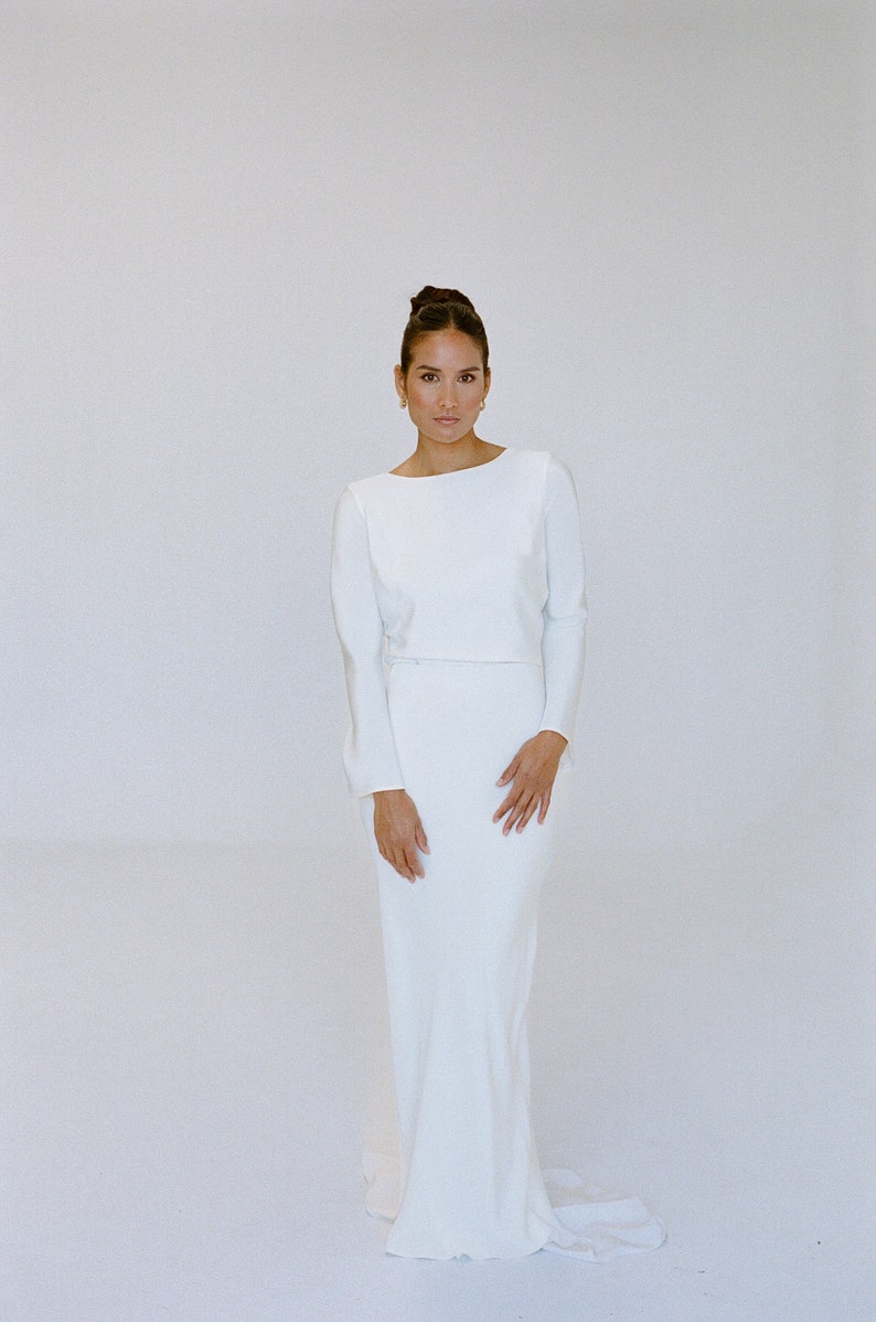 May include: A floor-length, ivory satin wedding dress with long sleeves and a fitted silhouette. The bodice is a cropped style, creating a defined waistline. The skirt flows smoothly to the floor, with a subtle train. The dress is simple and elegant, suitable for a modern bride.