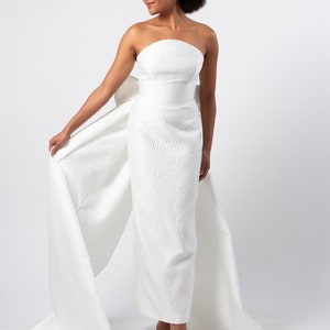May include: A white strapless gown with a large bow in the back. The gown has a textured fabric and a fitted silhouette.