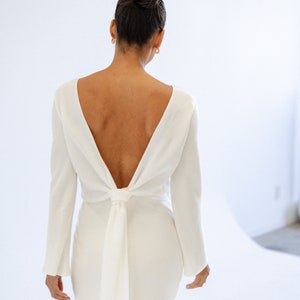 May include: A form-fitting, ivory white maxi dress with long sleeves and a deep V-back that ties at the waist. The dress is made of a smooth, flowing fabric and features a simple, elegant design.  The back features a unique knot detail. This minimalist wedding dress is perfect for a modern bride.