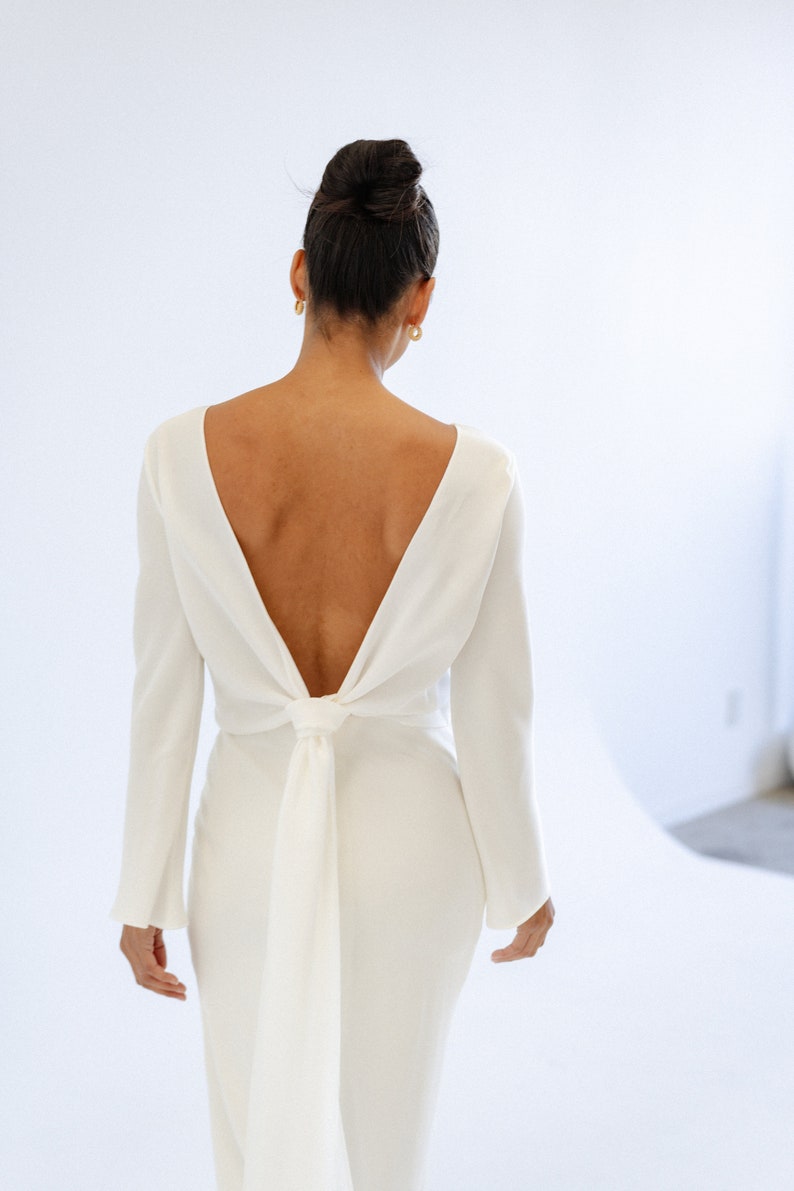 May include: A form-fitting, ivory white maxi dress with long sleeves and a deep V-back that ties at the waist. The dress is made of a smooth, flowing fabric and features a simple, elegant design.  The back features a unique knot detail. This minimalist wedding dress is perfect for a modern bride.