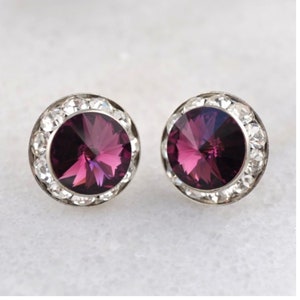 May include: A pair of silver stud earrings with a round, purple crystal surrounded by a halo of smaller, clear crystals.