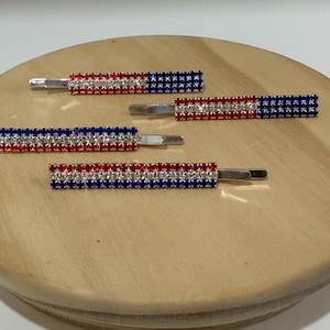 Puede incluir: Cuatro horquillas para el pelo con un diseño rojo, blanco y azul, dispuestas sobre una superficie de madera. Cada horquilla tiene forma rectangular con un clip de metal plateado. Las horquillas están decoradas con pequeños brillantes.