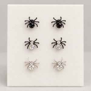May include: Three pairs of spider-shaped stud earrings arranged in a vertical column. The top pair is black with black crystals, the middle pair is black with clear crystals, and the bottom pair is silver-toned metal with clear crystals.