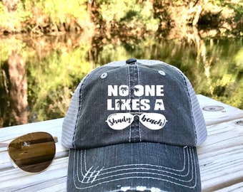 May include: A gray and white distressed trucker hat with the text "No one likes a shady beach" and a pair of sunglasses with palm trees in the lenses.