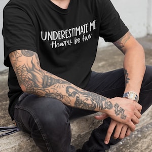 May include: A black t-shirt with white text that reads "UNDERESTIMATE ME that'll be fun".