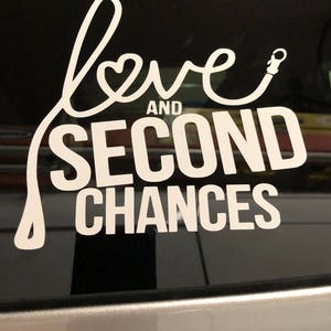 Op de afbeelding: Witte vinyl sticker met de tekst "love and second chances" in een cursief schrift. Het woord "love" wordt gevormd door een hartvorm en een riem met een clip.