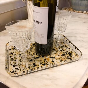May include: A clear tray with black and gold flecks, holding two clear stemmed glasses and a bottle of wine labeled "Del Atrio".