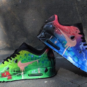 Puede incluir: Un par de zapatillas Nike Air Max 90 pintadas a mano. Una zapatilla est&aacute; pintada en tonos de verde, azul y rosa, mientras que la otra est&aacute; pintada en tonos de azul, rosa y negro. Ambas zapatillas presentan un dise&ntilde;o &uacute;nico de pintura salpicada.