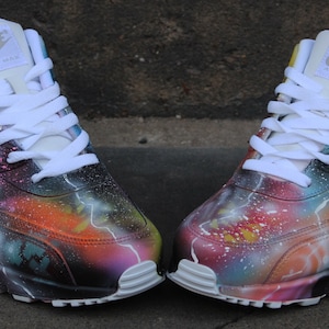 Op de afbeelding: Een paar witte Nike Air Max 90 sneakers met een galaxy-thema verfdesign. De sneakers hebben een kleurrijk spuitverfdesign met sterren, planeten en bliksemschichten. Het Nike-logo is zichtbaar aan de zijkant van elke schoen.
