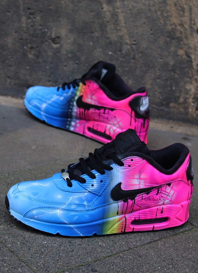 May include: A pair of custom-painted Nike Air Max 90 sneakers. One shoe is painted with a blue and pink gradient with lightning bolt accents, while the other shoe is painted with a pink and blue gradient with a black swoosh logo.