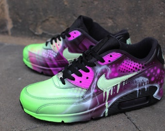 May include: A pair of custom-painted Nike Air Max 90 sneakers. One shoe is green with black and purple accents, while the other is purple with black and green accents. Both shoes feature a white swoosh logo and a black sole.
