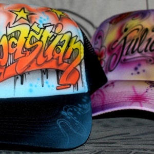 Peut inclure: Deux casquettes de camionneur personnalis&eacute;es. L'une est noire avec un filet blanc &agrave; l'arri&egrave;re et porte le nom "Sebastian" en lettres de style graffiti color&eacute;es. L'autre est rose avec un filet blanc &agrave; l'arri&egrave;re et porte le nom "Julia" en &eacute;criture cursive avec des motifs floraux.